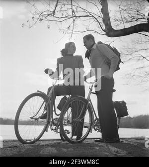 1940s couple on bicycles. A young couple is out on a tour on their bicycles on a sunny spring day. They have practical bags attached to the racks of the bicycles where they could transport the picnic food.  Sweden 1947. Photo Kristoffersson AB11-10 Stock Photo