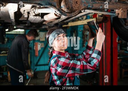 Portrait of a young beautiful female mechanic in uniform who conducts a car inspection using a diagnostic gadget. The car is on the lift. Stock Photo