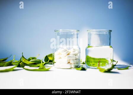 Two glass jars with pills on a blue background and a white table. Green leaves and white pills. Health theme. Stock Photo