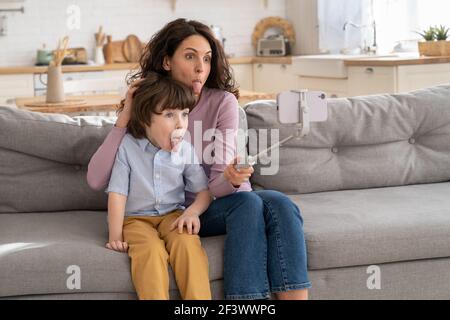 Mother and kid holding phone on selfie stick for video call showing tongue sitting on couch at home Stock Photo