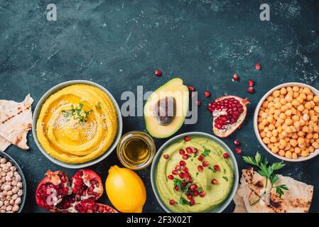 Ingredients for yellow hummus and green hummus - chickpea, tahini, olive oil, sesame seeds, pita, avocado, pomegranate on dark background. Middle Stock Photo
