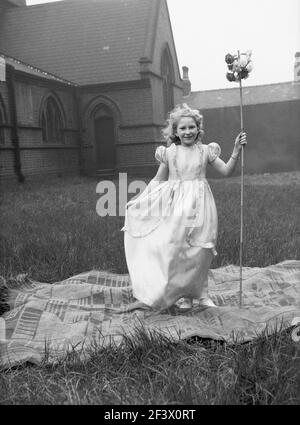 1958, historical, springtime and a young girl in her pretty parade costume and holding her flower stick, standing on a rug in the grounds of a church before taking part in the May day festival, Leeds, England, UK, May first,  commonly known as May Day, is traditonally a celebrated spring day, as well as a celebration of the roman goddess of flowers, Flora. Stock Photo