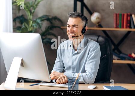 Online communication concept. Influential successful caucasian adult business man in formal wear and headset sits in his office communicates via video conference with colleagues using computer Stock Photo