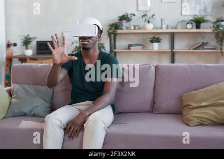 Young African man using VR goggles at home he sitting on sofa and gesturing Stock Photo