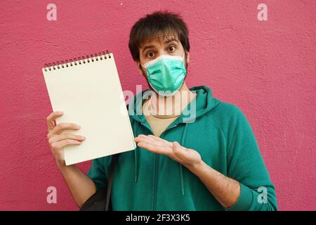 A surprised Hispanic male from Spain with a mask pointing at a notebook against pink wall - copy space Stock Photo