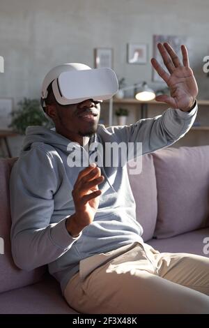 African young man in vr goggles gesturing while sitting on sofa during playing video game Stock Photo
