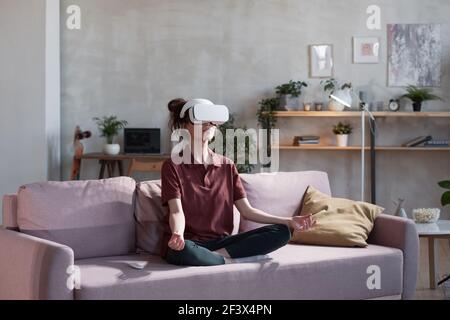 Young woman in vr glasses sitting on sofa in lotus position and meditating during virtual reality Stock Photo