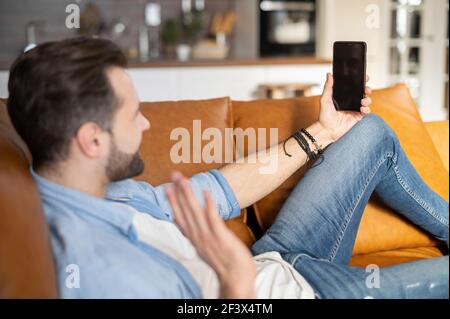 Positive young bearded hipster guy lying down on the couch and talking online on the mobile phone with a blank screen, video-calling with the family and coworkers, greeting friend with a high five Stock Photo