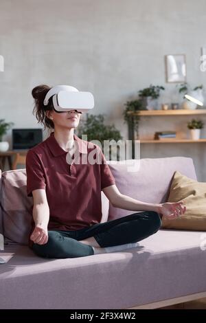 Young woman in vr headset sitting on sofa and meditating while watching yoga lesson in virtual reality Stock Photo