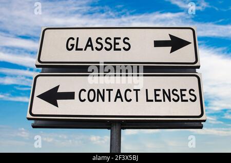 Glasses versus contact lenses road sign with two arrows on blue sky background. White two street sign with arrows on metal pole. Two way road sign wit Stock Photo