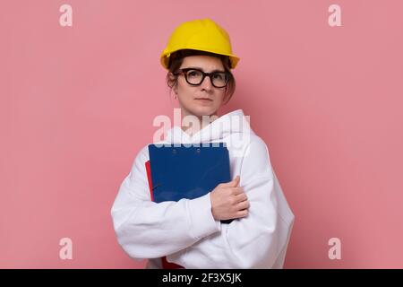 Young craftswoman in glasses and yellow helmet looking at camera. Studio shot on pink wall. Stock Photo