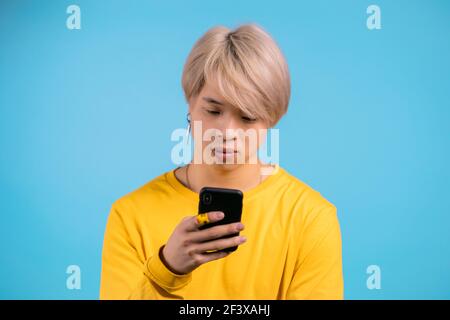 Handsome korean man using smartphone on blue studio wall. Blond guy in yellow outfit using modern technology - apps, social networks. Stock Photo