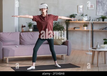 Young woman in VR glasses standing on exercise mat and exercising in the room Stock Photo