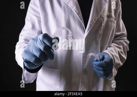 A dentist in a lab coat and gloves holding transparent mouthguards isolated on black background Stock Photo
