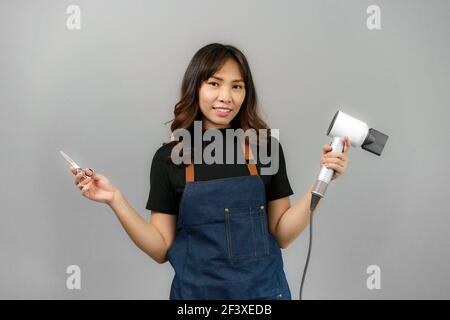 Portrait of Asian gorgeous cheerful young female professional hairdresser holding blowdryer smiling to the camera posing at her beauty studio.