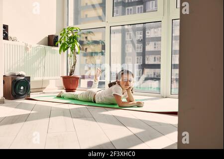 Beautiful little girl child resting on a yoga mat against the background of large windows at home