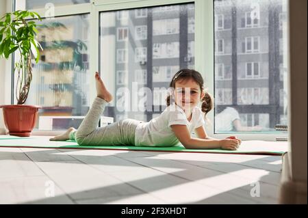 Adorable girl relaxing on an exercise mat and smiles while posing to camera against the background of large windows at home on a sunny day