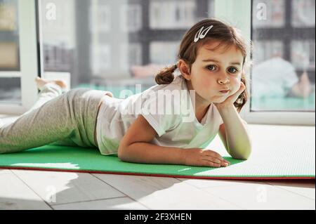 Adorable little girl relaxing on an exercise mat and posing to camera against the background of large windows at home on a sunny day