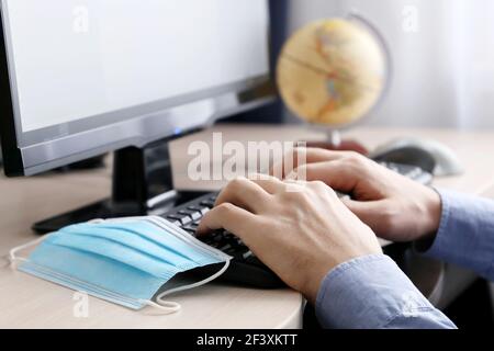 Man with removed protective mask typing at sitting at PC keyboard on globe background. Office work during coronavirus pandemic, travel agen Stock Photo