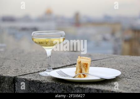 Glass of white sparkling wine and plate with sweet cake on the stone parapet on city background. Concept of travel, celebration, romantic dinner Stock Photo