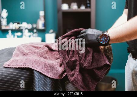 Hands of barber wearing gloves applying hot towel to male client's face to moisturize beard hairs before old fashioned straight razor shave. Stock Photo