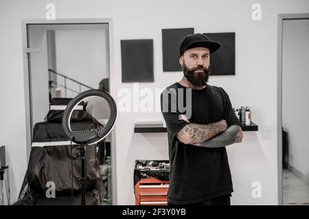 Young male tattoo artist with beard looking positive and happy standing and smiling in workshop place. Stock Photo