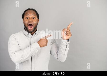 Surprised with unbelievable deal or new product an African-American man points fingers away at empty copy-space, amazed multiethnic guy with opened mouth attracts attention, isolated on grey Stock Photo