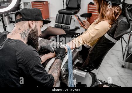 Male tattoo master tattooing female client checking phone. Tattoo machine and lamp. Workplace artist. Creating picture on hand with it in salon. Stock Photo