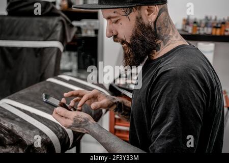 Young male tattoo artist with beard checking his phone sitting on couch in workshop place. Stock Photo