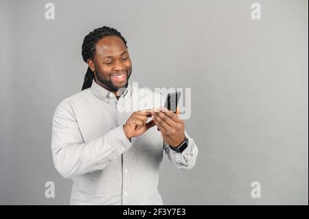 Joyful African-American man with using smartphone isolated on grey, a black guy spends leisure in social networks, chatting online, shopping, using new mobile app, playing game on the phone Stock Photo