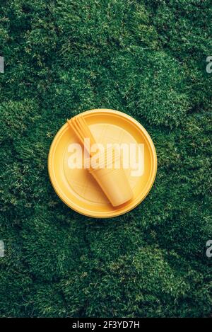 Environmental pollution. Disposable plastic tableware - plastic plates, forks, spoons, knives. Yellow plastic tableware on green grass, moss Stock Photo
