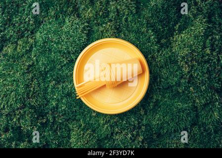 Yellow plastic tableware on green grass, moss background. Top view. Copy space. Environmental pollution. Ban single use plastic. Disposable plastic Stock Photo