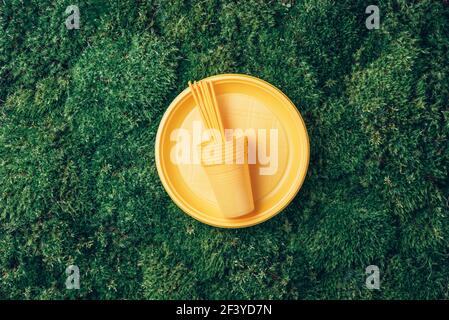 Yellow plastic tableware on green grass, moss background. Top view. Copy space. Environmental pollution. Ban single use plastic. Disposable plastic Stock Photo
