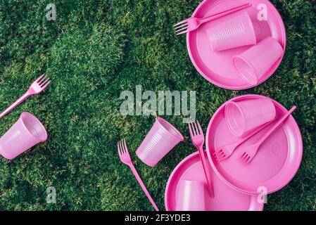 Environmental pollution. Disposable plastic tableware - plastic plates, forks, spoons, knives. Pink plastic tableware on green grass, moss background Stock Photo
