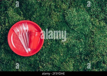 Red plastic tableware on green grass, moss background. Top view. Copy space. Environmental pollution. Ban single use plastic. Disposable plastic Stock Photo