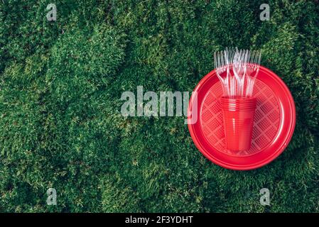 Environmental pollution. Disposable plastic tableware - plastic plates, forks, spoons, knives. Red plastic tableware on green grass, moss background Stock Photo