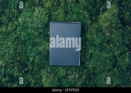 Craft notebook, black sketch book on fresh spring green grass, moss background. Copy space. Top view. Wild nature, ecology concept. Summer forest Stock Photo