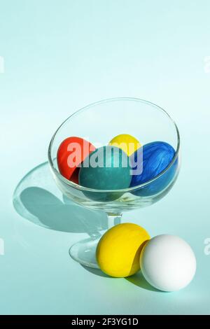 Hand-painted colorful Easter eggs in a transparent glass bowl on a light blue background, vertical frame, copy space, close-up. Happy Easter concept. Stock Photo