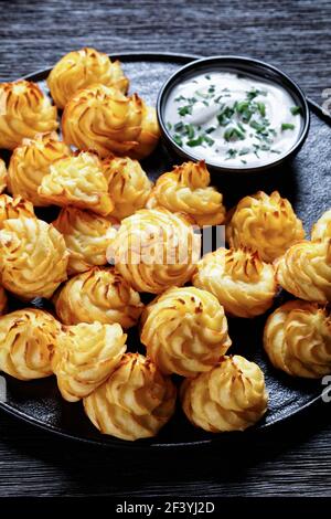 Duchess potatoes, mashed potatoes piped in decorative swirls, browned in an oven served with white sauce on a black plate on a wooden table Stock Photo