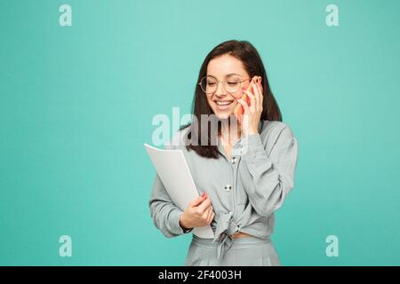 Photo of cute business lady in glasses holds papers and talking on phone. Wears grey shirt, isolated turquoise color background Stock Photo