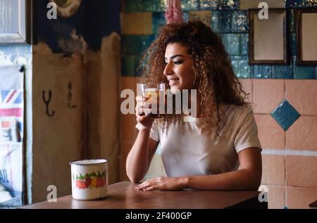 Young arabic woman with thoughtful gesture sitting in a beautiful bar with vintage decoration. Arab girl in casual clothes drinking a soda. Stock Photo