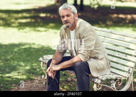 Pensive mature man sitting on bench in an urban park.. Portrait of a pensive mature man sitting on a bench in an urban park. Senior male with white hair and beard wearing casual clothes. Stock Photo