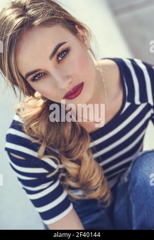Blonde woman, model of fashion, possing in urban background.. Close-up portrait of blonde woman, model of fashion, possing in urban background. Beautiful young girl wearing striped t-shirt and blue jeans in the street. Pretty russian female with pigtail. Stock Photo