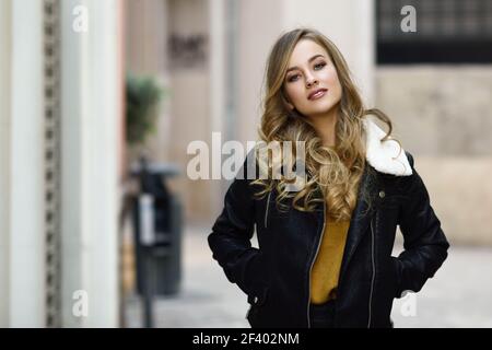 Blonde woman in urban background. Beautiful young girl wearing black leather jacket and mini skirt standing in the street. Pretty russian female with long wavy hair hairstyle and blue eyes. Stock Photo
