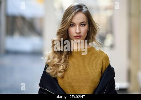 Blonde woman in urban background. Beautiful young girl wearing black leather jacket and mini skirt standing in the street. Pretty russian female with long wavy hair hairstyle and blue eyes. Stock Photo