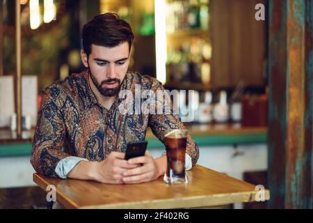 Young bearded man wearing casual clothes looking at his smartphone in a modern pub. Guy with beard and modern hairstyle drinking a cola. Stock Photo