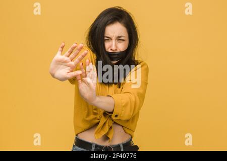 Concept photo of female victim with her mouth taped up, holds her head. Wears yellow shirt, isolated yellow color background Stock Photo