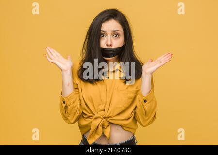 Concept photo of female victim with her mouth taped up. Wears yellow shirt, isolated yellow color background Stock Photo