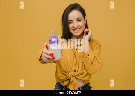 Photo of cute female holds baby bootle of milk. Wears yellow shirt, isolated yellow color background Stock Photo