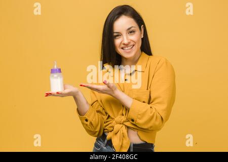 Photo of cute female holds baby bootle of milk. Wears yellow shirt, isolated yellow color background Stock Photo